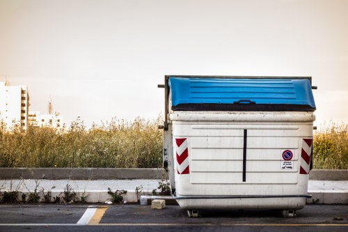 A large commercial waste skip positioned on a paved area adjacent to a curb, with a blue hinged lid on top and white sides reinforced with red and white striped reflective markings. The skip appears to be made of sturdy metal or heavy-duty plastic, with a slightly textured surface and some visible dirt or minor scuffs indicating previous use. It is situated in an outdoor setting near an overgrown grassy area with a few weeds along the edge of the pavement. Behind the skip, there is a stretch of low shrubbery and blurred distant buildings under a clear or slightly overcast sky, suggesting a quiet urban or suburban environment. The image is captured during daylight hours, with natural lighting casting soft shadows. The scene reflects routine rubbish collection and disposal activities typical around Herne Hill and surrounding London suburbs, aligning with services provided by Commercial Waste Herne Hill for efficient rubbish removal.