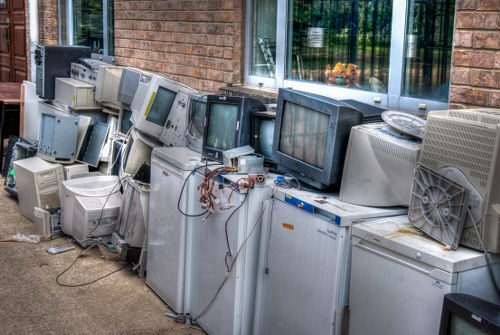 A row of discarded, vintage computer monitors and electronic appliances arranged along a brick building exterior, positioned on a paved surface. The monitors vary in size and shape, with several featuring plastic casings in shades of grey, beige, and black. Some screens are turned off, showing blank dark displays, while others have visible buttons and controls on their front panels. A few devices have tangled wires and cables hanging loosely, indicating they are no longer in use. Behind the electronic waste, there are large glass windows with reflections of greenery and a partly visible interior, suggesting the area may be adjacent to an office or residential building in Herne Hill. The scene reflects a typical electronic waste collection or disposal area, emphasizing the need for professional rubbish removal services like those provided by Commercial Waste Herne Hill for responsible recycling and disposal of electronic items.