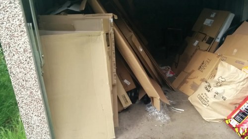 A small pile of stored rubbish inside a garage or storage area, consisting of various cardboard boxes and packaging materials. The cardboard boxes are of different sizes, some flattened, with printed labels and logos visible. The boxes and packaging are positioned haphazardly, leaning against the garage wall, with some stacked and others scattered around the floor. In the background, a dark interior space is partially visible, suggesting a cluttered storage environment. Outside the garage, a section of paved driveway or pathway can be seen with a small strip of grass at the edge of the frame. The scene appears typical of rubbish accumulation awaiting removal or disposal, reflecting the kind of waste management services provided by Commercial Waste Herne Hill to commercial clients in the local area near Herne Hill, London.